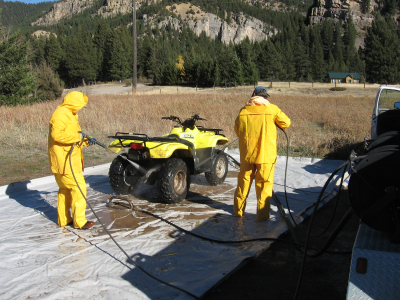 Two people in yellow rain gear spraying a dirty four wheeler. Brown grass and green conifer trees are in the background.
